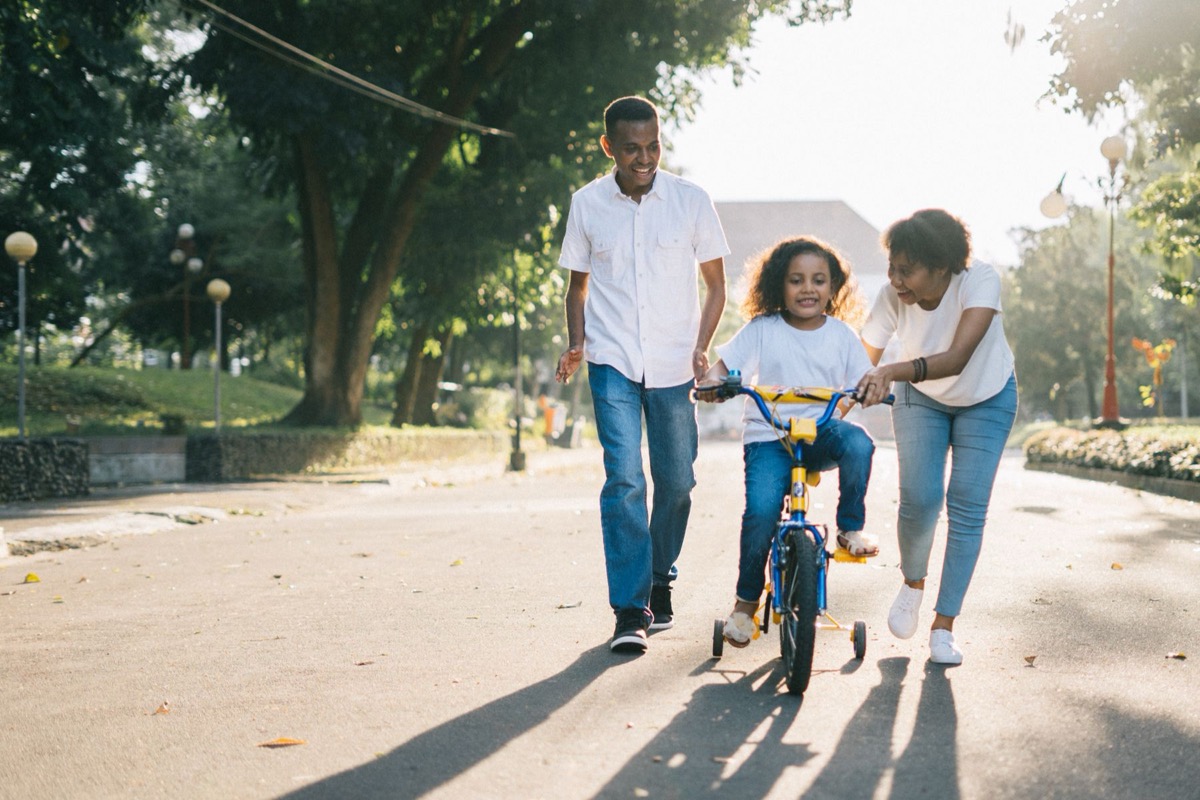 A parent and grandparent running alongside a child learning to ride a bicycle in a park at golden hour.